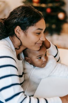 Side view of positive ethnic mother in casual clothes sitting with baby on hands and looking away in apartment against Christmas tree